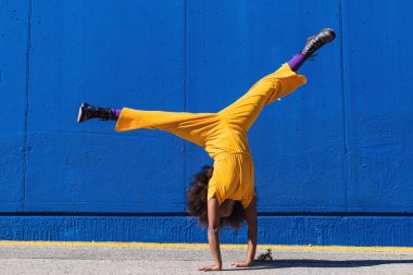Açık sarı tulumlu Afro-Amerikan bir genç kız caddede mavi duvara karşı amuda kalkıyor.