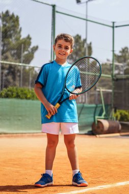 Spor kıyafetli, pis bir tenis kortunda tenis oynayan küçük bir çocuğun portresi.