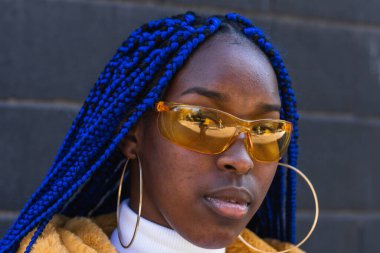 Mavi örgülü, sarı ceketli, kendine güvenen Afro-Amerikan kadın sokakta dikilip kameraya bakıyor.