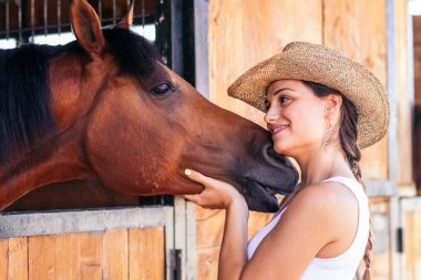 Genç bir kadın ahırda atına bakıyor. At sakin ve rahat görünürken, bakıcı ona dikkat ve özen gösterir..