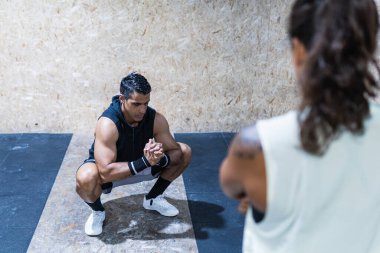 Ciddi kaslı İspanyol erkek sporcu. Spor giyim kuşamında. Hafif spor salonunda antrenman yaparken duvara yakın çömelmiş ve kimliği belirsiz bir kadın.