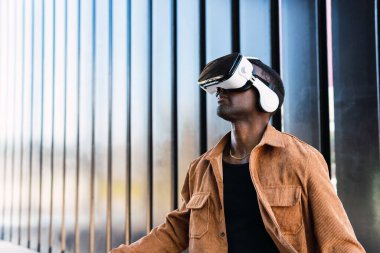 VR gözlüklü Afro-Amerikalı bir erkek siber uzayı keşfederken modern binanın yanında metal duvarlı bir caddede duruyor.