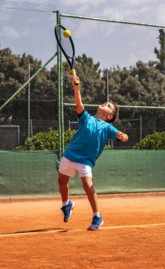 Spor giyim kuşağında tenis oynayan küçük çocuk tenis kortunda elinden gelenin en iyisini yapmaya çalışıyor.