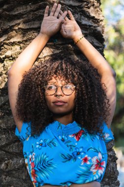 Palmiye gövdesine yaslanmış kıvırcık saçlı ve yaz günü gözlerini kaçıran zarif siyah kadın.