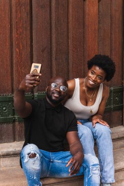 Neşeli Afro-Amerikalı çift merdivenlerde oturmuş akıllı telefondan selfie çekiyorlar.