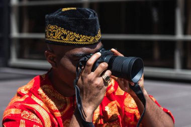 Anonymous black man in traditional red clothes and kufi taking picture on professional photo camera in sunny day in city street clipart