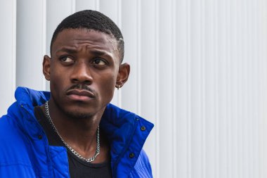 Kendinden emin Afrikalı Amerikalı erkek model canlı mavi bir ceket giyiyor. Sokakta metal bir duvarın üzerinde dikilip başka tarafa bakıyor.