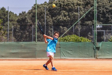 Spor giyim kuşağında tenis oynayan küçük çocuk tenis kortunda elinden gelenin en iyisini yapmaya çalışıyor.