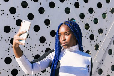 Kararlı Afro-Amerikan kadın mavi örgülü şehir ortamında duruyor ve akıllı telefonun selfie kamerasıyla fotoğraf çekiyor.