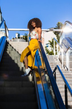 Kıvırcık saçlı Afro-Amerikan kadın, güneşli bir yaz gününde yürüyen merdivende dikiliyor.