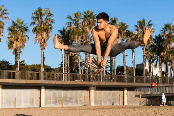 Güçlü genç Asyalı sporcu çıplak gövdesiyle yoğun spor yaparken kumlu sahilde geniş bacaklı atlama egzersizi yapıyor.