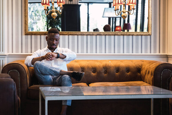 Serious bearded African American businessman in trendy outfit sitting on couch and checking time on wristwatch while waiting for meeting in stylish cafe