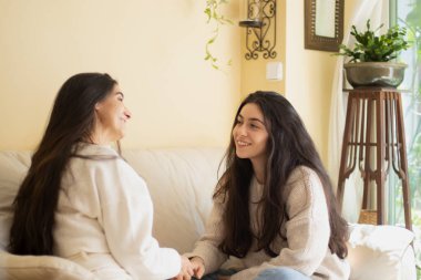 Gülümseyen genç bir kadın annesiyle evdeki kanepede konuşuyor..