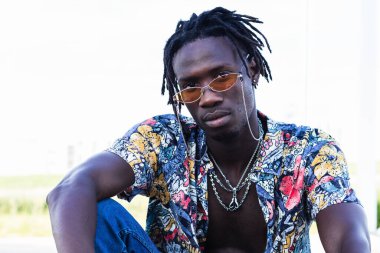 Alçak açılı, renkli gömlekli ve kot pantolonlu ciddi bir Afro-Amerikan erkeği şehir caddesinde oturuyor ve kameraya bakıyor.