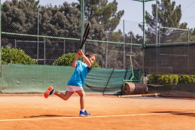 Spor giyim kuşağında tenis oynayan küçük çocuk tenis kortunda elinden gelenin en iyisini yapmaya çalışıyor.