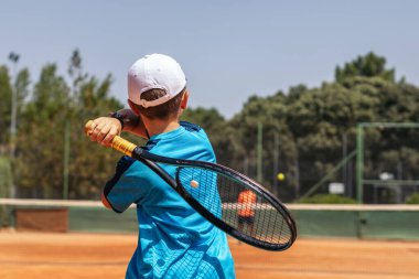 Spor giyim ve şapkalı küçük bir çocuk tenis kortunda tenis oynarken elinden gelenin en iyisini yapmaya çalışıyor.