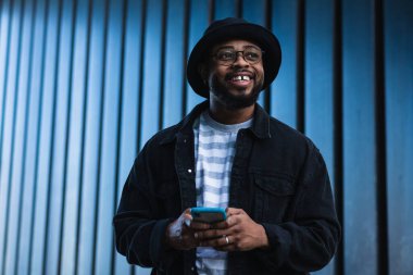 Neşeli yetişkin sakallı Afro-Amerikan erkek. Günlük kıyafet ve şapka giymiş. Duvarın karşısında duruyor ve akıllı telefondan mesaj yolluyor.