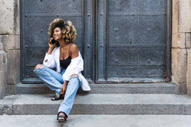 Pozitif Afro-Amerikalı kadın, günlük kıyafetleriyle oturmuş cep telefonuyla konuşuyor ve etrafa bakıp gülümsüyor.