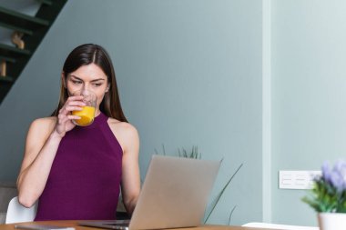 Modern netbook kullanarak, bir bardak taze portakal suyuyla masada oturan çekici, güler yüzlü bir kadın.