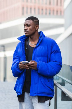 Sıcak ceketli Afro-Amerikan erkek bina yakınında duruyor ve akıllı telefondan sosyal medyada mesajlaşıyor.