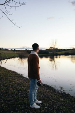 Gün batımında bir gölün manzarasına bakarken kamerasını tutan bir fotoğrafçının arka görüntüsü.