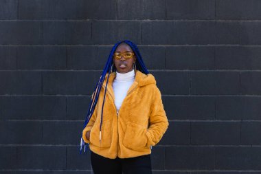 Mavi örgülü, sarı ceketli, kendine güvenen Afro-Amerikan kadın sokakta dikilip kameraya bakıyor.