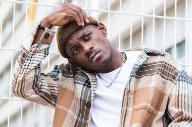 Şık gömlekli, şapkalı, çitlere yaslanmış modern şehir sokaklarına bakan genç bir zenci.