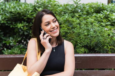 Parkta bankta oturup başka tarafa bakarken cep telefonuyla konuşan mutlu bir kadın.