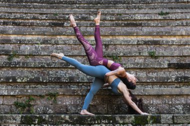 Sıska kadınlar spor kıyafetleriyle birlikte akro yoga yapıyor ve taş merdivenlerde vücutlarını esnetiyor.