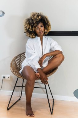 Çekici Afro-Amerikan kadın vücudu kıvırcık saçlı evde şık hasır koltukta otururken kameraya bakıyor.