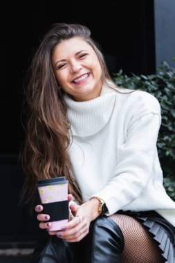 Büyüleyici, rüya gibi bir kadın elinde bir fincan sıcak kahveyle kameraya bakıyor ve parkta boş vakitlerin tadını çıkarırken gülümsüyor.