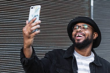 Şapkalı ve gözlüklü neşeli yetişkin Afrikalı Amerikalı hippi sokakta metal duvarın yanında dururken cep telefonundan selfie çekiyor.