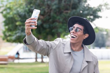 Neşeli etnik stil erkek modaya uygun kıyafetler ve şapkalar giyiyor geniş bir gülümseme ve modern binanın yanında cep telefonuyla kendi portresini çekiyor.