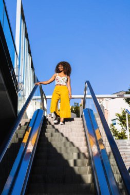 Kıvırcık saçlı Afro-Amerikan kadın, güneşli bir yaz gününde yürüyen merdivende dikiliyor.