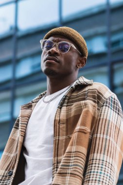 Moda desenli tişörtlü genç siyahi adam modern şehrin sokaklarında takılırken kaşlarını çatıyor ve kameraya bakıyor.