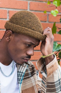 Kareli gömlekli ve modaya uygun şapkalı genç Afro-Amerikan adam sokağın karşısında tuğla duvara bakıyor ve kaşlarını çatıyor.