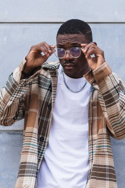 Moda desenli gömlekli Afro-Amerikalı adam şehir sokaklarında duvara yaslanırken gözlük takıyor.