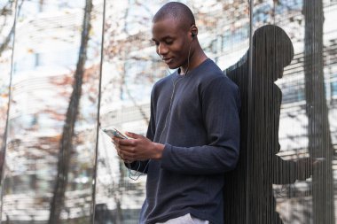Neşeli Afrikalı Amerikalı adam, kulaklıkla müzik dinlerken cam duvarın yanında cep telefonu kullanıyor.