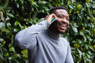 Yeşil çalıların yanında telefonla konuşurken gülen sıradan giyinmiş Afro-Amerikan bir erkek.