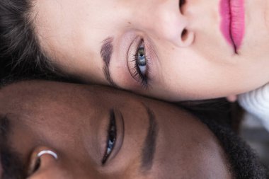 Yukarıdan, çok ırklı genç çiftin yakın planından. Yüz yüze yatıp, birlikte romantik vakit geçirirken kameraya bakıyorlar.