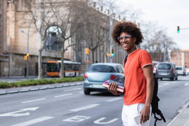 Kıvırcık saçlı ve gözlüklü, üniversiteden önce sırt çantası ve kitaplarla yol kenarında otobüs bekleyen şık Afro-Amerikan erkek.