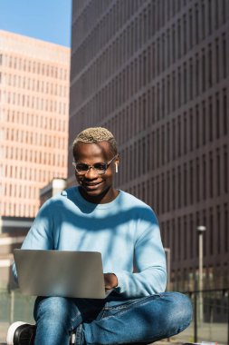 Kendine güvenen, modaya uygun kıyafetli, gözlüklü, gözlüklü Afro-Amerikan bir adam. Şehir parkında uzaktan kumandayla yazı yazıyor. Elinde kahve ve defteriyle bankta oturuyor.