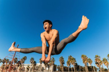 Güçlü genç Asyalı sporcu çıplak gövdesiyle geniş bacaklı atlama egzersizi yaparken yoğun fitness antrenmanı sırasında kumsalda çığlık atıyor.