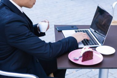Klas takım elbiseli Asyalı bir girişimci kafede dizüstü bilgisayarla masada oturuyor ve iş sırasında kahve molası verirken tatlı kek yiyor.