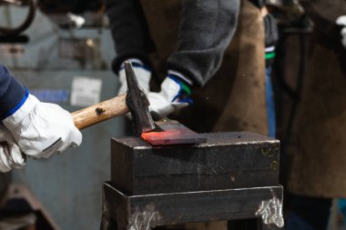 Çekiçle tanınmayan erkek ustalar örse yerleştirilmiş sıcak metal parçalarına vuruyor.