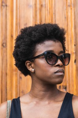 Genç, mutsuz, kıvırcık saçlı, güneş gözlüğü takan, tahta duvara yaslanmış, düşünceli bir şekilde başka tarafa bakan Afro-Amerikan kadın.