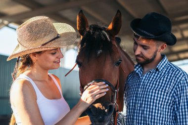 Kovboy tarzı şapkaları ve çizmeleri olan genç bir adam ve kadın kırsal alanda atlarıyla birlikte yürüyorlar..