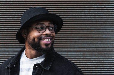 Gözlüklü, gözlüklü, modaya uygun kıyafet ve şapka takan parlak bir şekilde gülümseyen ve kepenkli duvara bakarken yüzünü çeviren mutlu Afro-Amerikan sakallı adam.