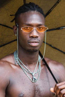 Ciddi bir Afro-Amerikan genci çıplak gövdesi ve rastaları olan, modaya uygun güneş gözlüğü takan, elinde şemsiyesi açık metal zincirler olan ve kameraya bakan.