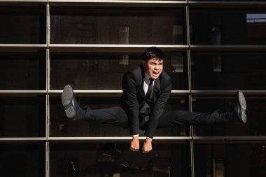 Resmi takım elbiseli Asyalı girişimci bağırıyor şehir merkezindeki cam binanın arka planında yükseğe zıplıyor ve başka tarafa bakıyor.